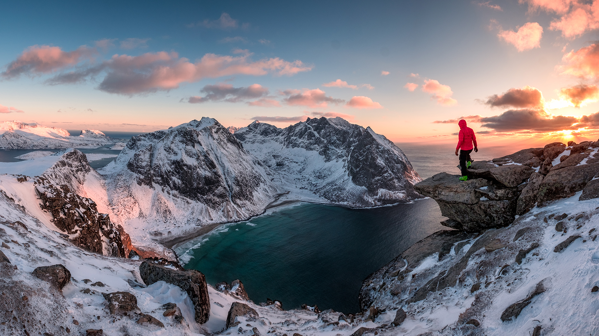 En person i rød jakke står på toppen av et snødekt fjell og skuer utover fjell og fjord.